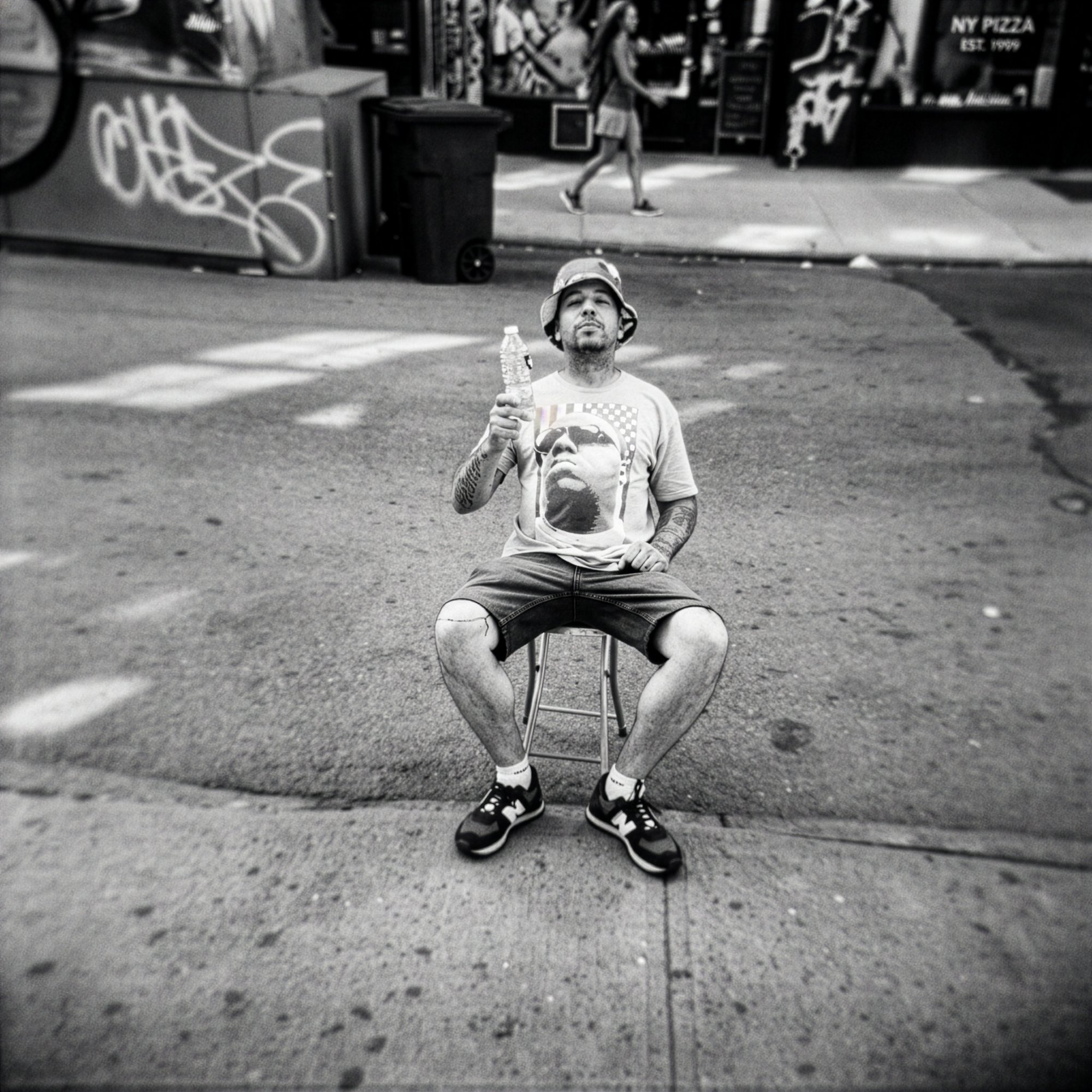 Young man on a folding stool in the middle of a SoHo street, Notorious B.I.G. tee, bucket hat, water bottle raised, B&W.